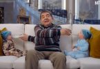 Rowan Atkinson sits on a sofa feeding two babies with milk bottles in opposite directions in a scene from Netflix’s Man vs. Baby.