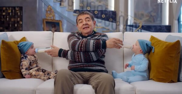 Rowan Atkinson sits on a sofa feeding two babies with milk bottles in opposite directions in a scene from Netflix’s Man vs. Baby.