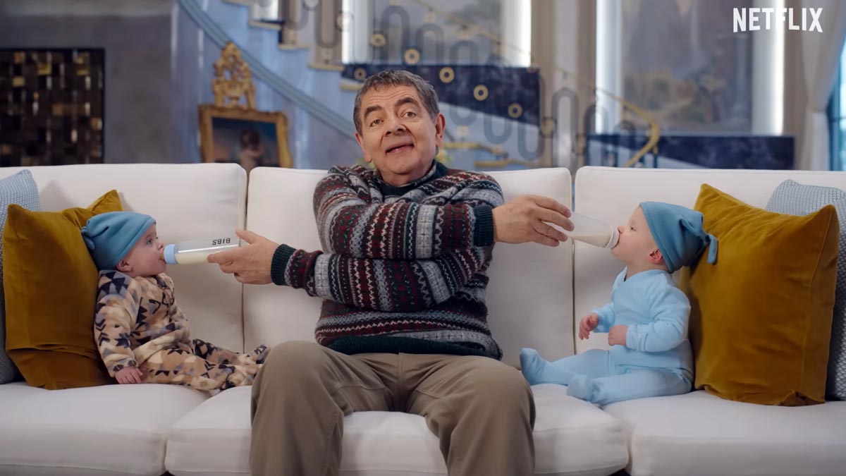 Rowan Atkinson sits on a sofa feeding two babies with milk bottles in opposite directions in a scene from Netflix’s Man vs. Baby.