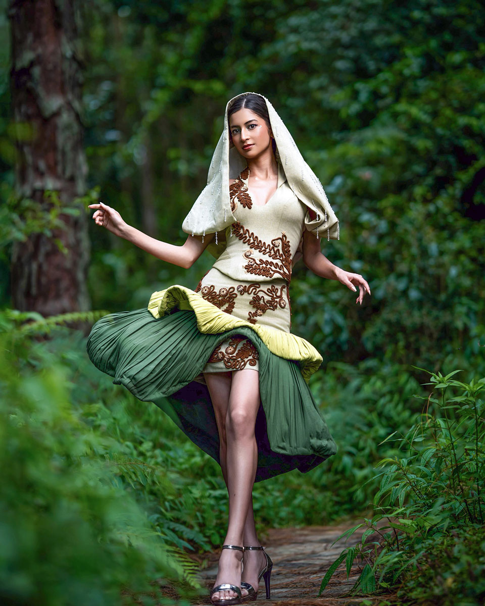 Model in a pale green dress with raised brown botanical motifs, a hood-like veil and a sculptural dark green drape, photographed among trees.