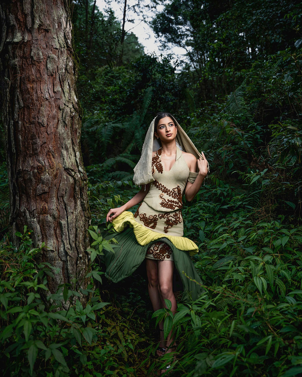 Model in a pale green dress with raised brown botanical motifs, a hood-like veil and a sculptural dark green drape, photographed among trees.