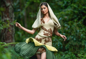 Model in a pale green dress with raised brown botanical motifs, a hood-like veil and a sculptural dark green drape, photographed among trees.