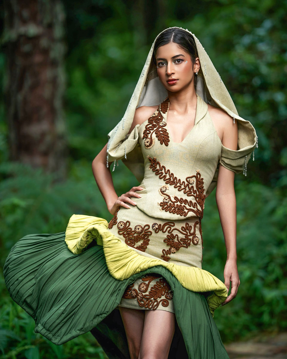 Model in a pale green dress with raised brown botanical motifs, a hood-like veil and a sculptural dark green drape, photographed among trees.