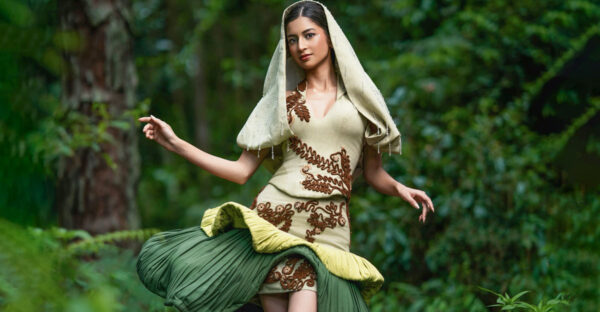 Model in a pale green dress with raised brown botanical motifs, a hood-like veil and a sculptural dark green drape, photographed among trees.