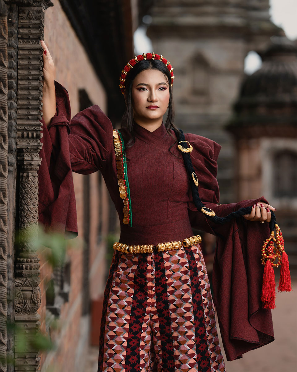 Model in a burgundy couture outfit with gathered sleeves, patterned wide-legged garment, gold-toned ornaments and a decorated long braid before carved Nepali architecture.