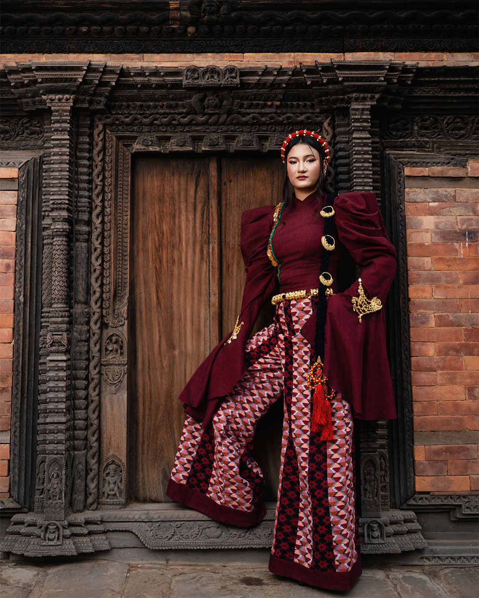 Model in a burgundy couture outfit with gathered sleeves, patterned wide-legged garment, gold-toned ornaments and a decorated long braid before carved Nepali architecture.
