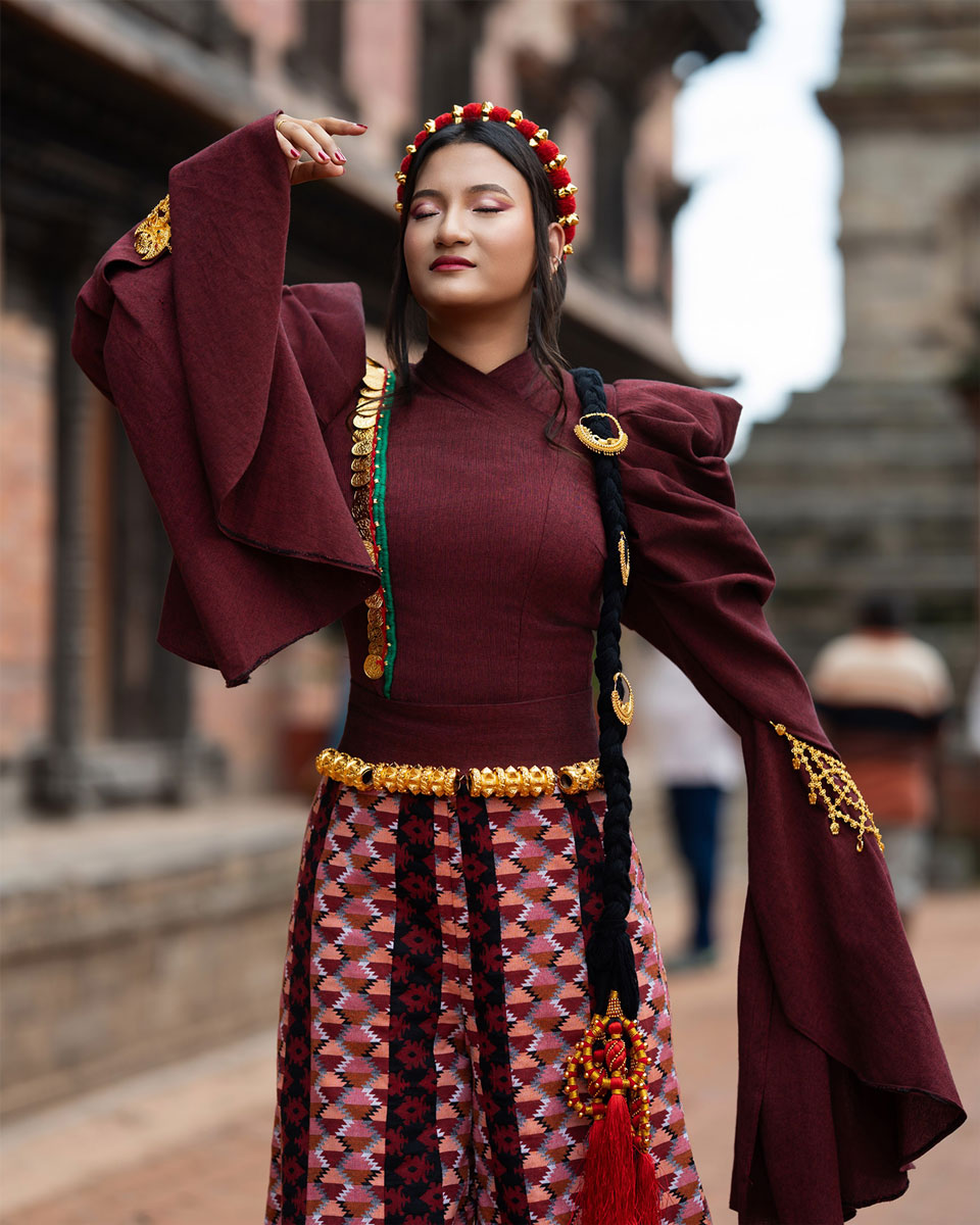 Model in a burgundy couture outfit with gathered sleeves, patterned wide-legged garment, gold-toned ornaments and a decorated long braid before carved Nepali architecture.