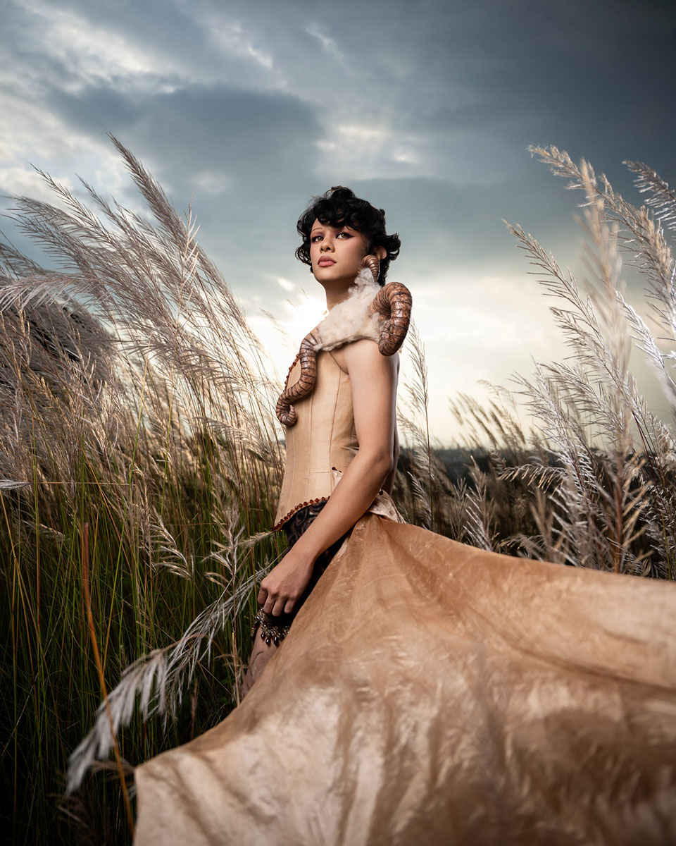 Model in a pale fitted corset and sweeping skirt with wool and curled horn-inspired shoulder pieces, photographed among tall grasses.