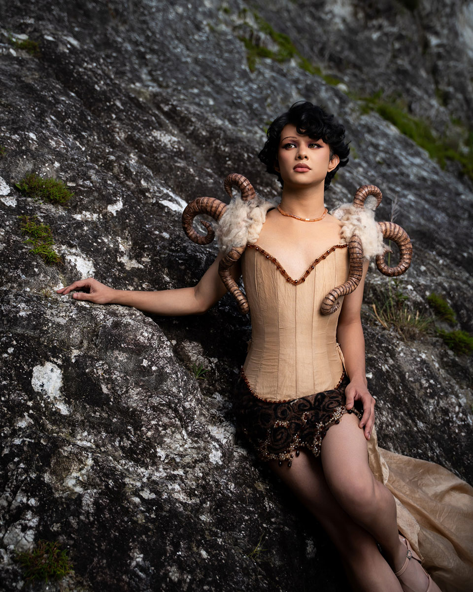 Model in a pale fitted corset and sweeping skirt with wool and curled horn-inspired shoulder pieces, photographed among tall grasses.