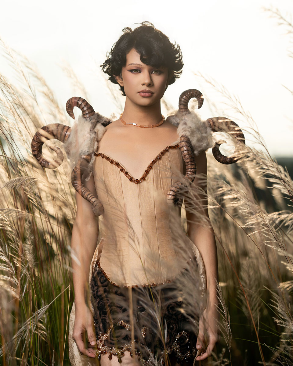 Model in a pale fitted corset and sweeping skirt with wool and curled horn-inspired shoulder pieces, photographed among tall grasses.