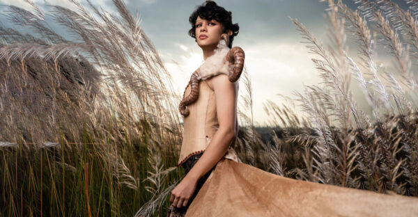 Model in a pale fitted corset and sweeping skirt with wool and curled horn-inspired shoulder pieces, photographed among tall grasses.