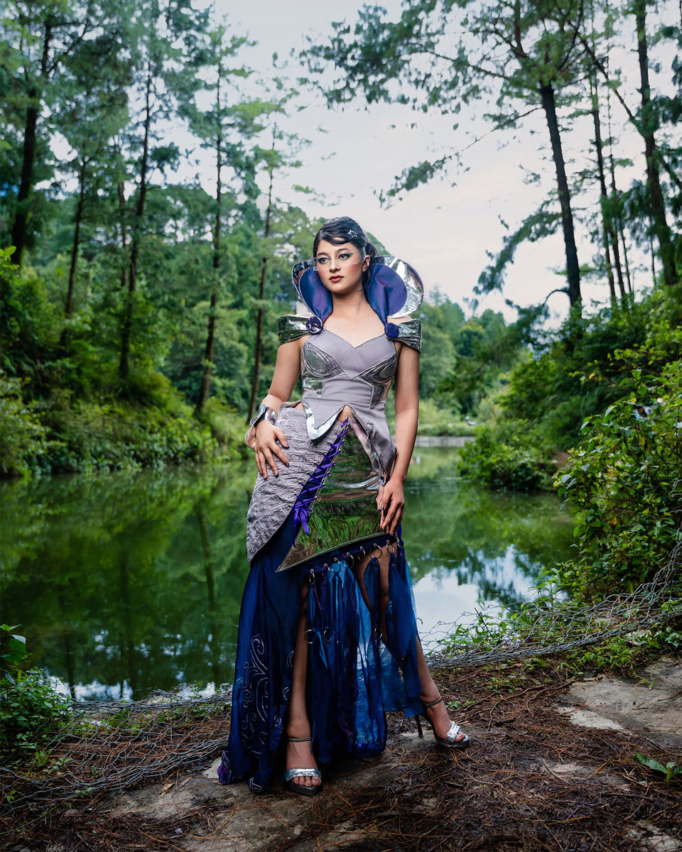 Model wearing a futuristic couture look with reflective silver structure, purplish-blue flowing fabric and a clear visor beside a wooded pond.