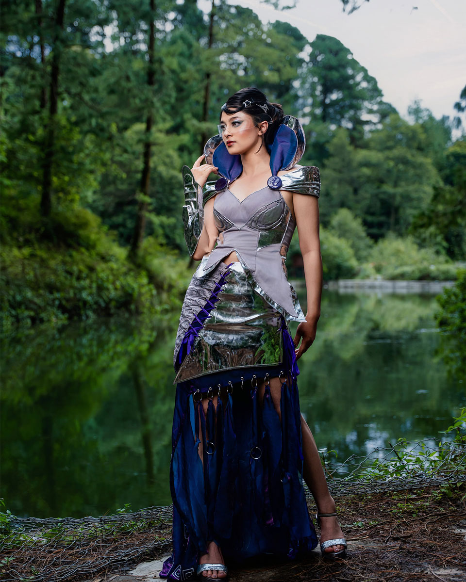 Model wearing a futuristic couture look with reflective silver structure, purplish-blue flowing fabric and a clear visor beside a wooded pond.