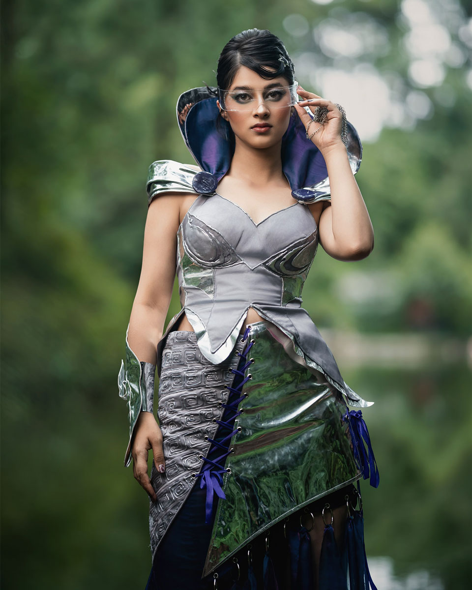 Model wearing a futuristic couture look with reflective silver structure, purplish-blue flowing fabric and a clear visor beside a wooded pond.