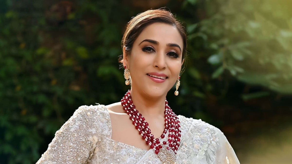 Nepali actress Niruta Singh smiling in an embellished silver saree, layered red beaded necklace and dangling earrings against a leafy green background.