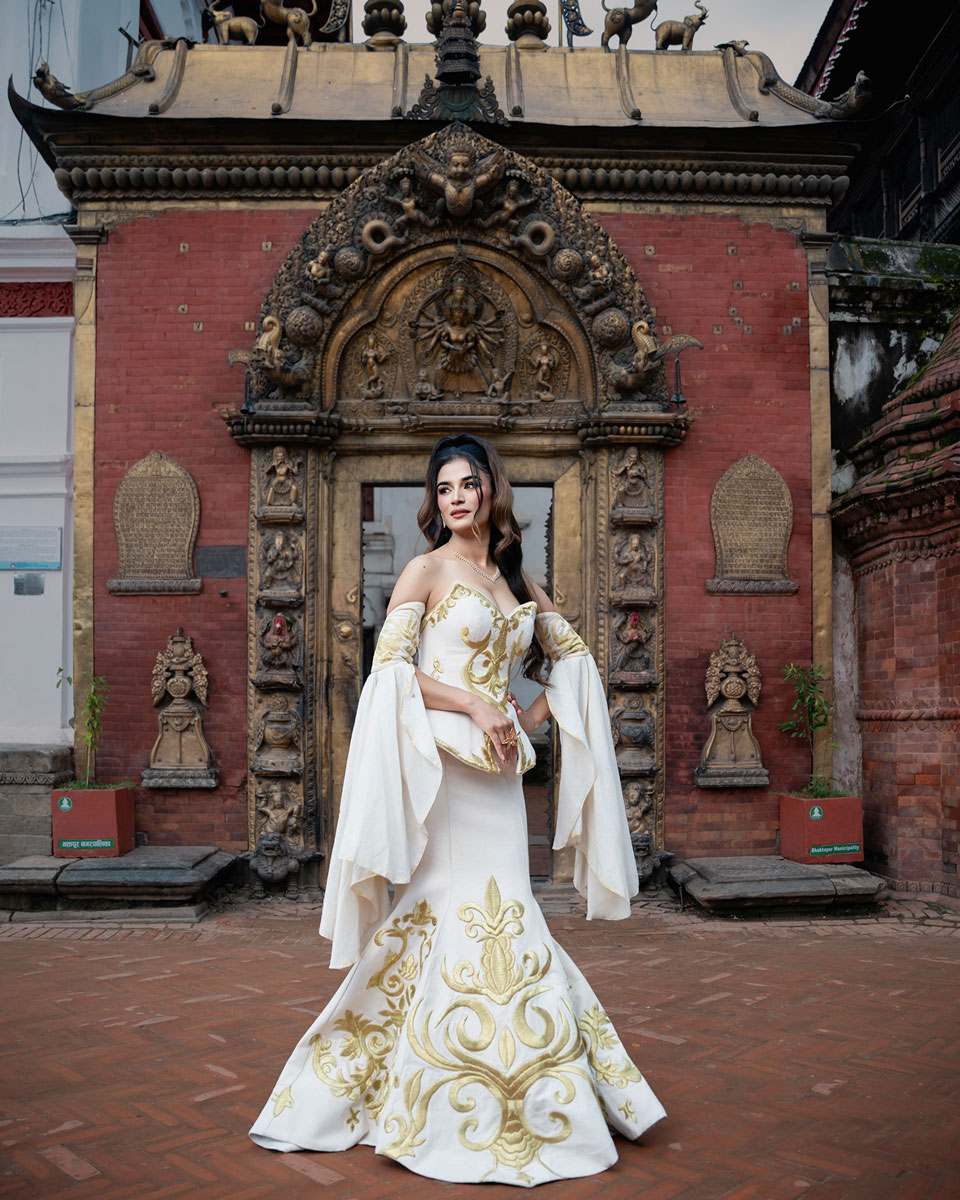 Model wearing an ivory mermaid gown with gold motifs, a sculpted corset and long flowing sleeves before a carved gateway.