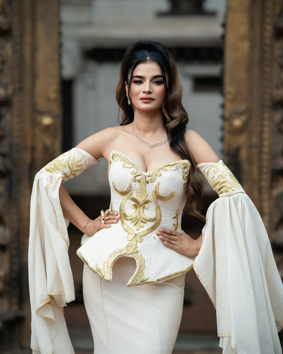 Model wearing an ivory mermaid gown with gold motifs, a sculpted corset and long flowing sleeves before a carved gateway.