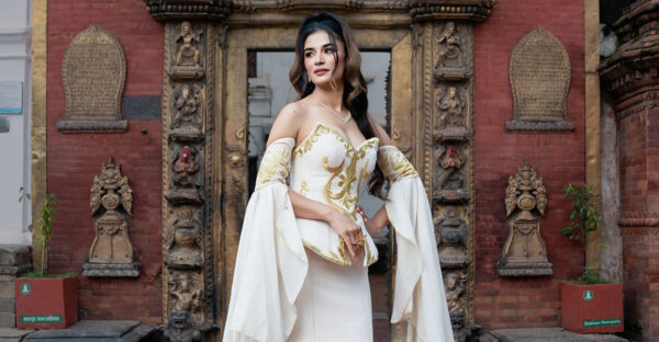 Model wearing an ivory mermaid gown with gold motifs, a sculpted corset and long flowing sleeves before a carved gateway.