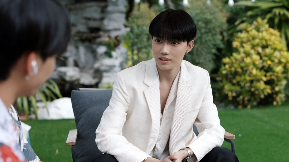 Siddhartha Tamang wearing a white blazer, seated opposite another character in a garden scene from Sawasdee Love.