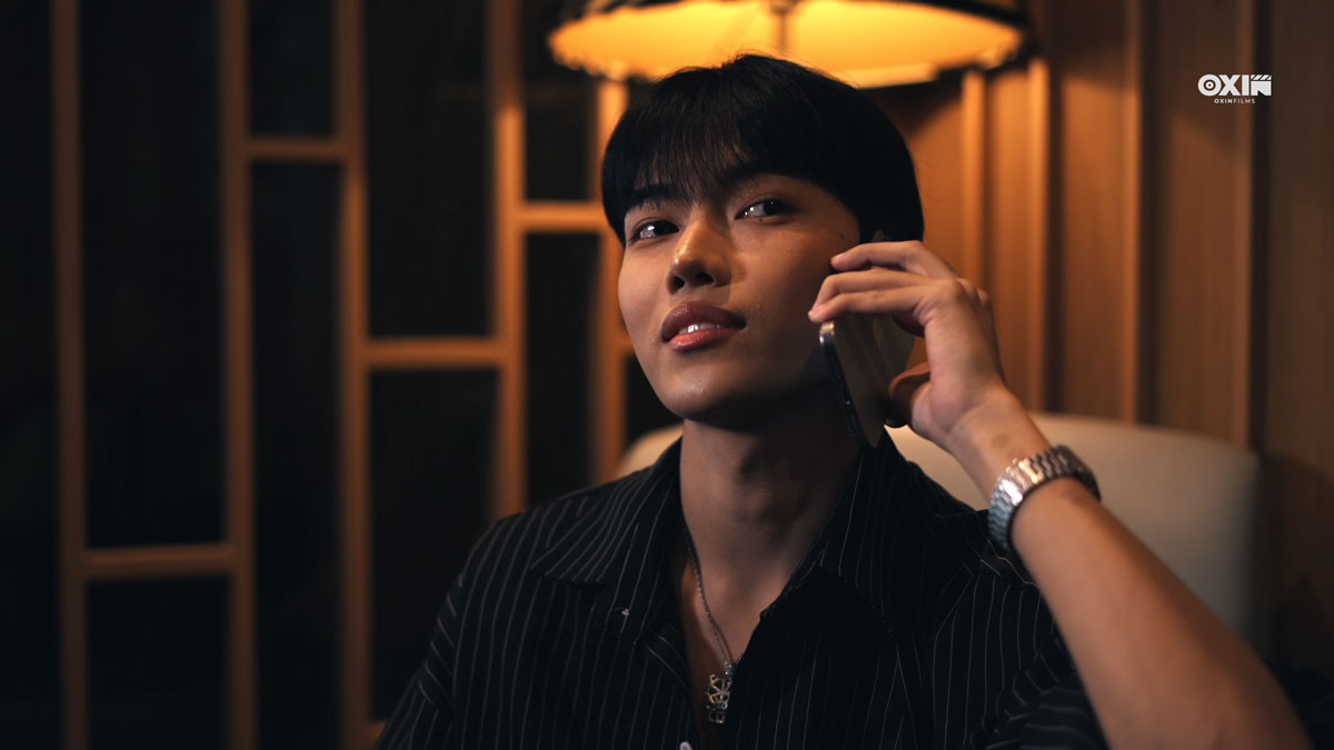 Siddhartha Tamang holding a phone to his ear, wearing a black pinstriped shirt in a warmly lit scene from Sawasdee Love.