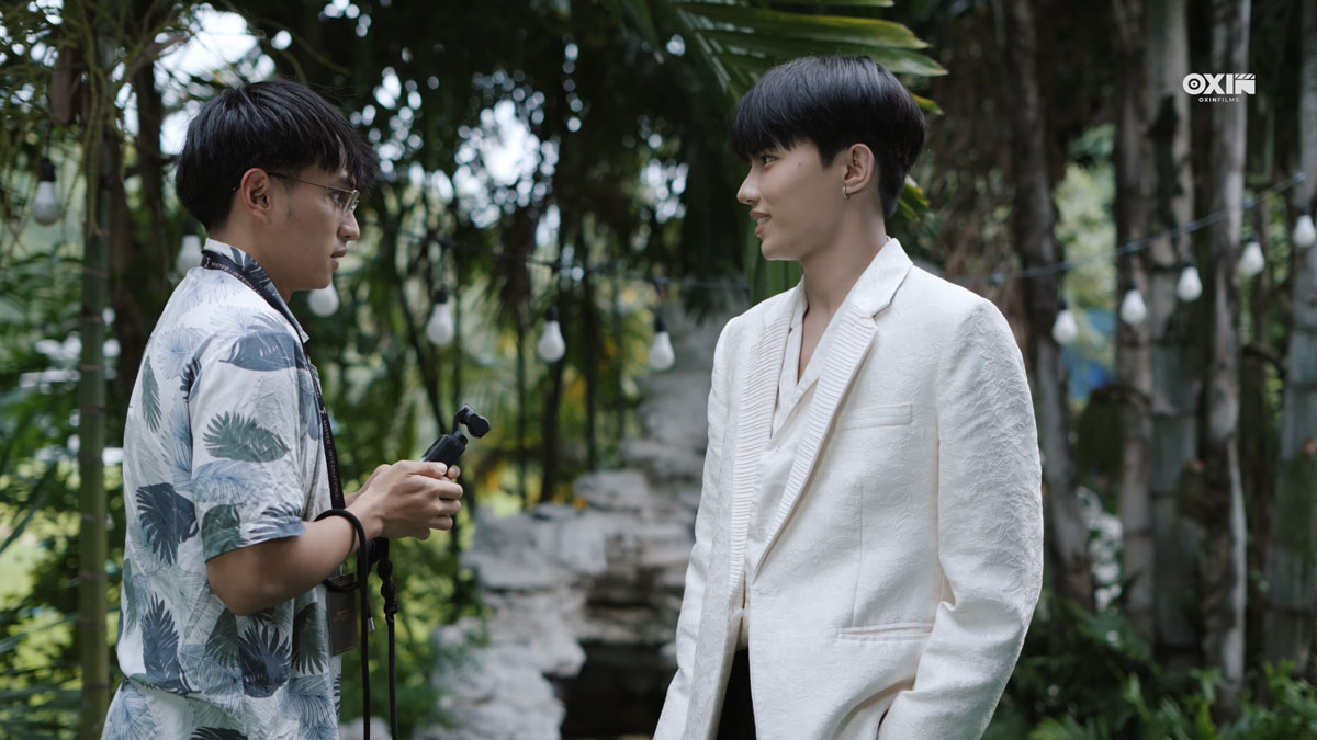 Siddhartha Tamang, right, in a white blazer, facing a man with glasses holding a small camera in a garden scene from Sawasdee Love.