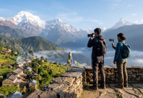 AI-generated image of two travel creators photographing and filming terraced hills, a Nepali village and snowy Himalayan peaks from a stone viewpoint.