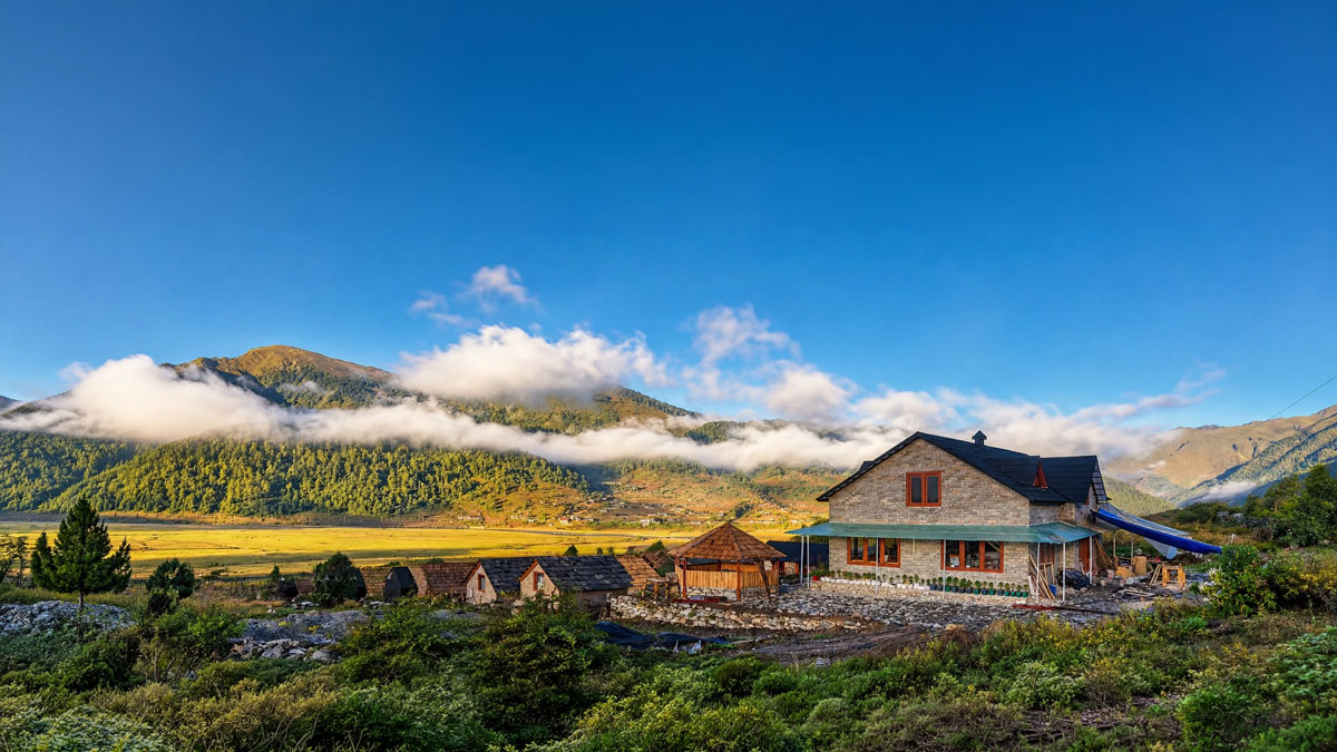 Panoramic view of SizzEcoCamp in Dhorpatan, showing mountain cottages, a stone-finished building, green meadows, forested hills and low clouds.